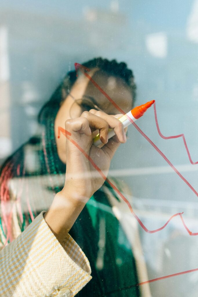 pexels-photo-7173047-7173047 Woman in a Beige Coat Writing on a Glass Panel Using a Whiteboard Marker